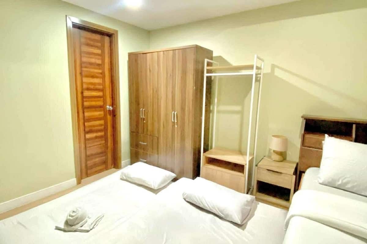 Habitación de hotel con dos camas, un armario de madera, cabina de ducha de vidrio y una mesita de noche con lámpara.