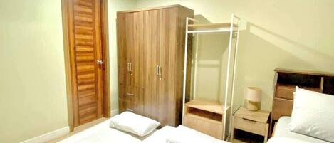 A hotel room with two beds, a wooden wardrobe, a glass shower enclosure, and a bedside table with a lamp.