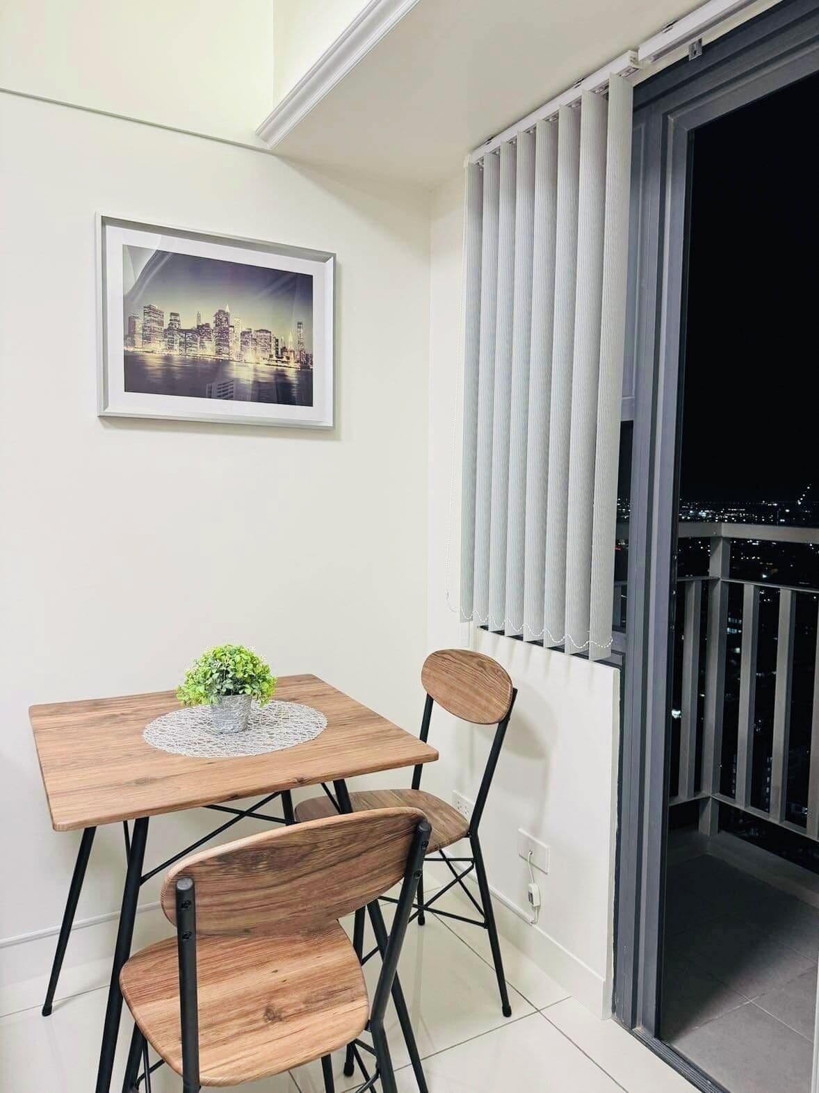 Un comedor con una mesa de madera y dos sillas, un cuadro en la pared con una vista urbana, y una vista de la ciudad a través de una ventana con persianas.