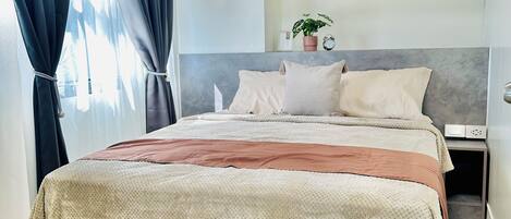 A modern bedroom with a large bed, a headboard with storage, a bedside table, and a window with curtains.