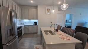 Una cocina moderna con armarios blancos, un refrigerador de acero inoxidable, un horno empotrado y una isla grande con pileta. Hay dos lámparas colgantes sobre la isla.