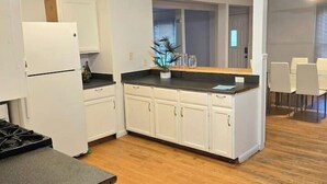 A modern kitchen with white cabinets, a black countertop, and a wooden floor. There is a refrigerator, a stove, and a dining area with chairs.