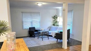 A living room with a wooden table, a sofa, and a coffee table. There is a window with blinds, a lamp, and a plant.