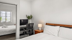 A hotel room with a bed, a desk with a computer, a chair, and a nightstand with a lamp.