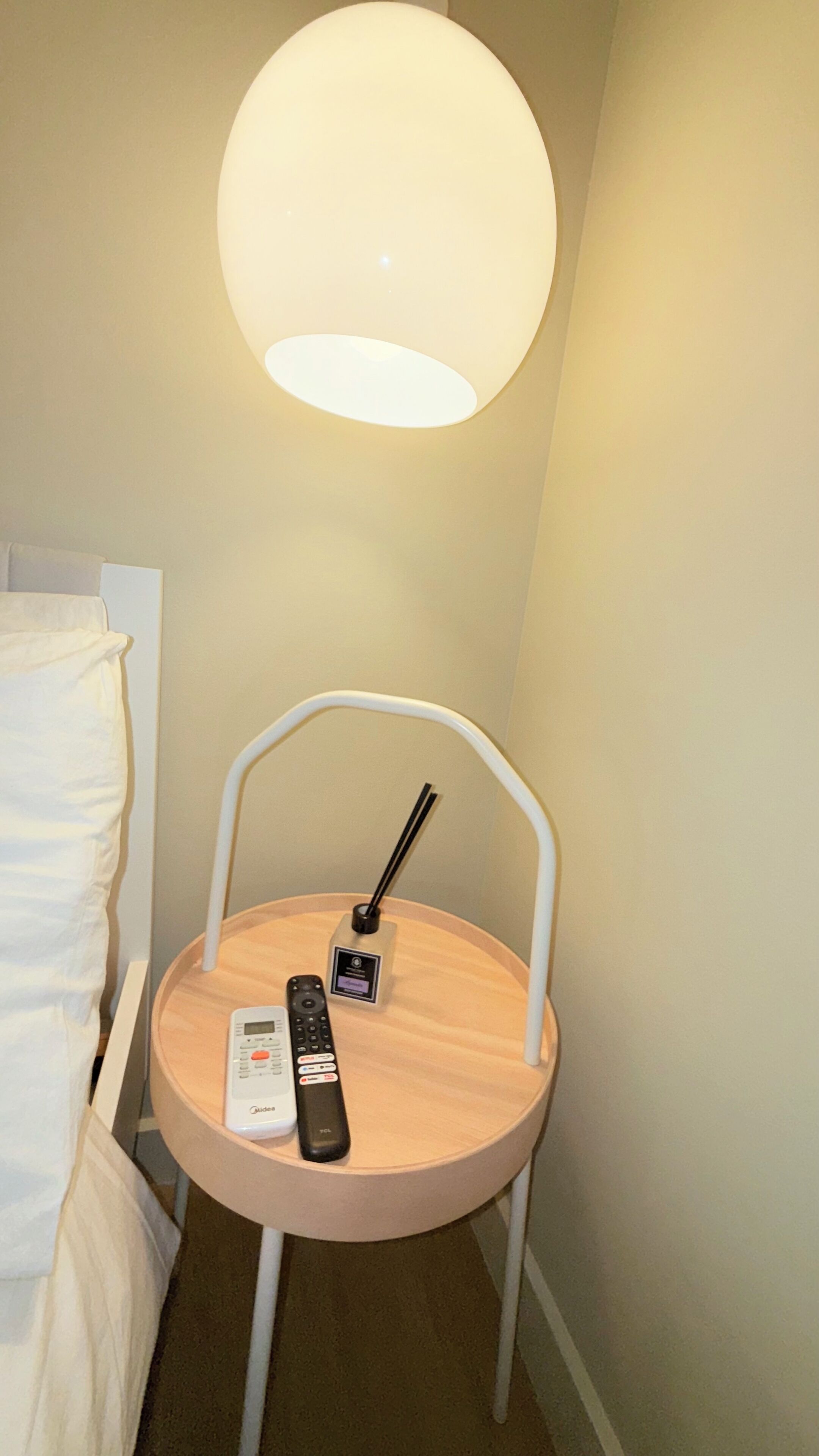 Une petite table ronde avec une télécommande et un multiprise, un lit avec du linge de lit blanc et un grand luminaire au plafond.