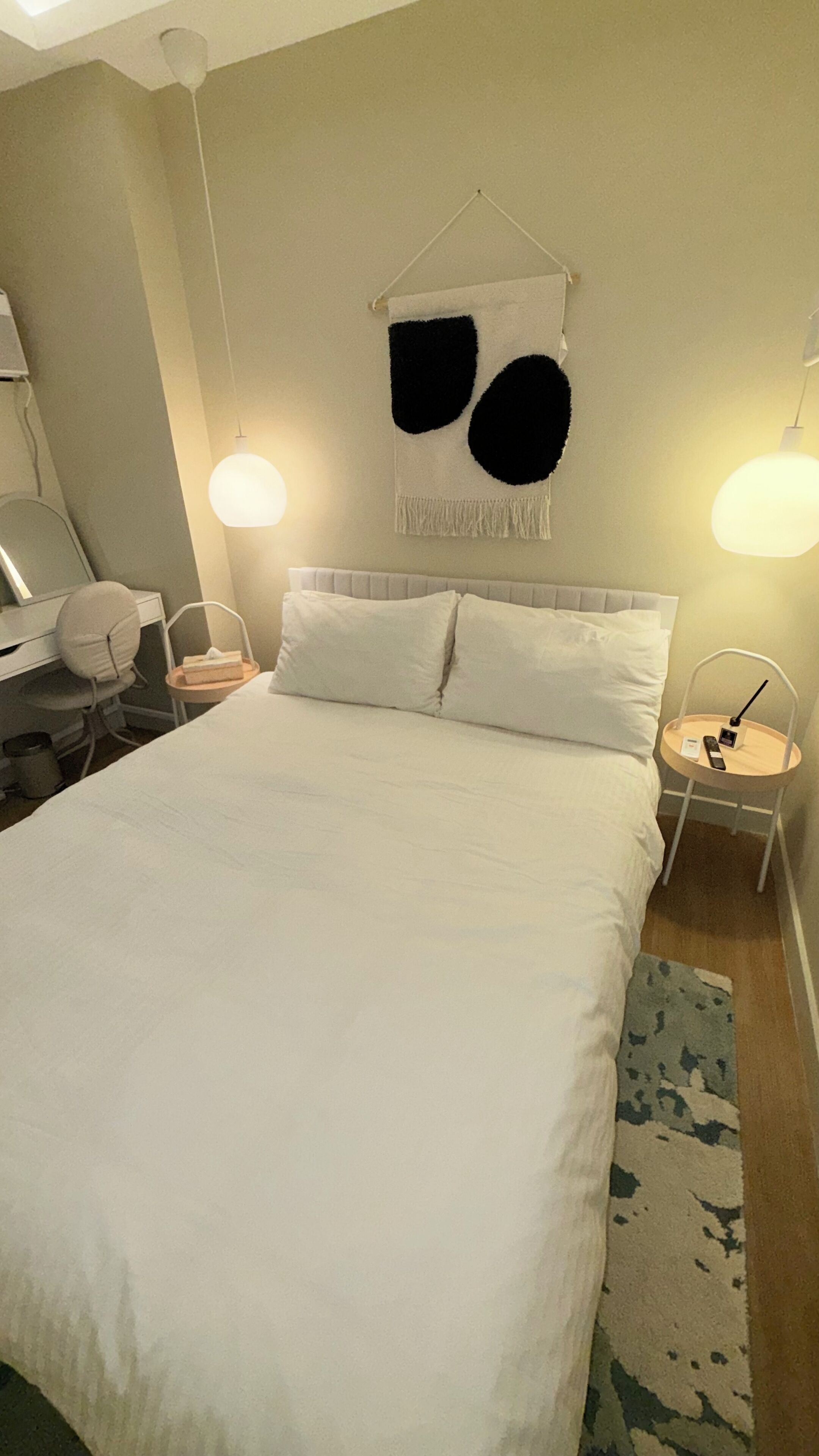 Een net opgemaakt bed met witte lakens, een hangend abstract kunstwerk en twee bedlampjes.