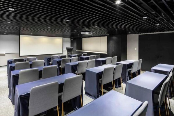 Un salón de conferencias con filas de mesas y sillas, una pantalla de proyector y una pantalla de presentación.