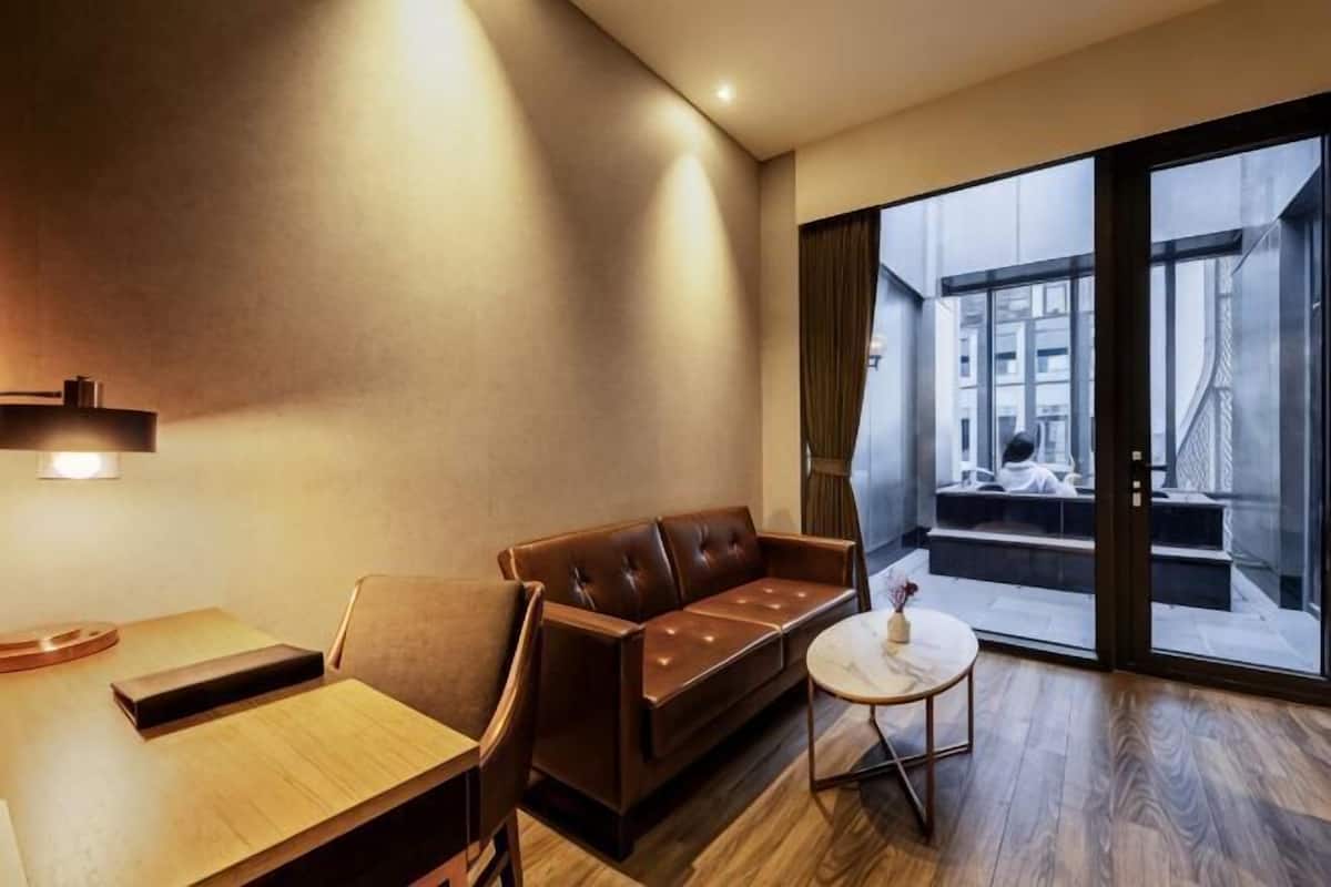 Una habitación de hotel moderna con piso de madera, un sofá de cuero, una pequeña mesa redonda y un amplio ventanal corredero que da a un balcón exterior.