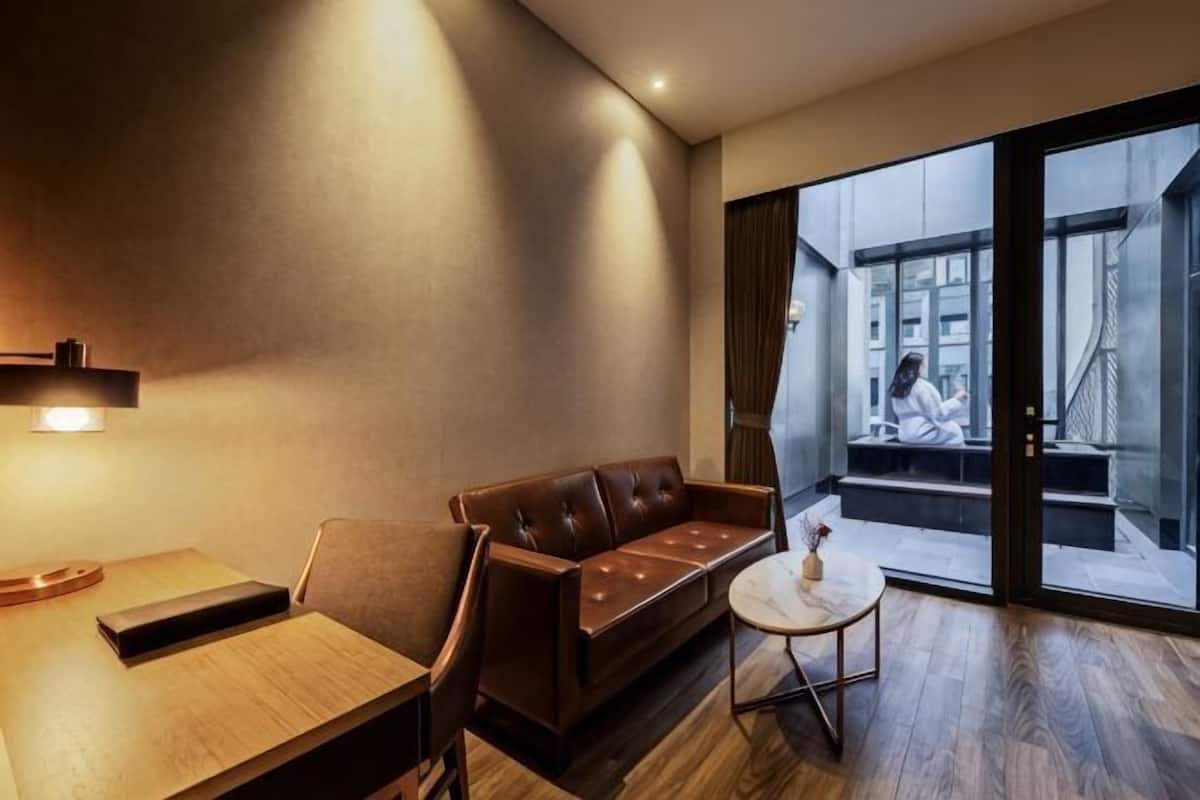 Una habitación de hotel moderna con un sofá de cuero, una pequeña mesa redonda y vistas a un paisaje urbano a través de puertas correderas amplias.