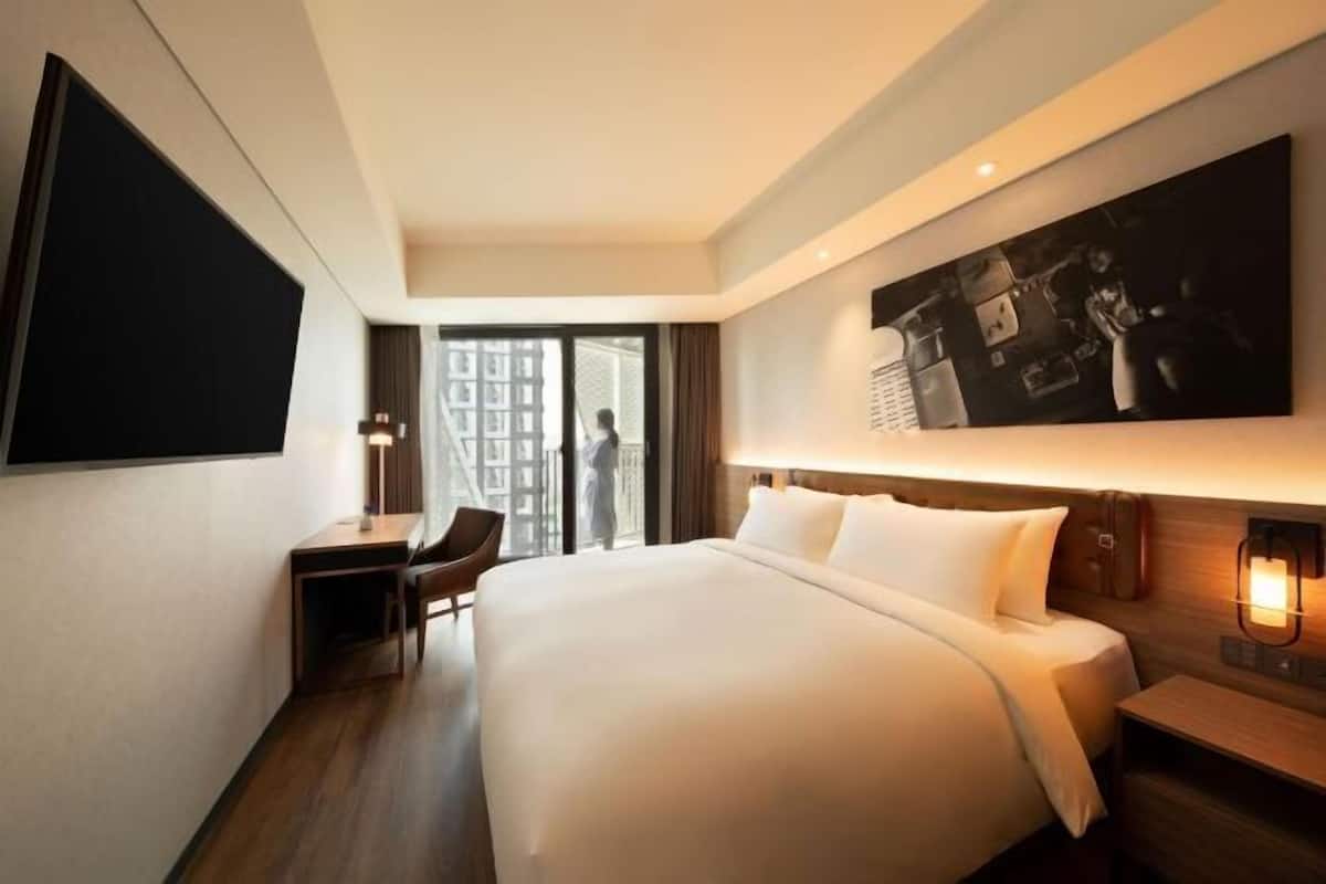 Habitación de hotel con una cama grande, un escritorio, una silla, un televisor fijado a la pared y un ventanal amplio.