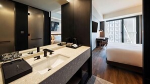 A modern hotel bathroom with a large sink, a mirror, and a bed with a view of the city.