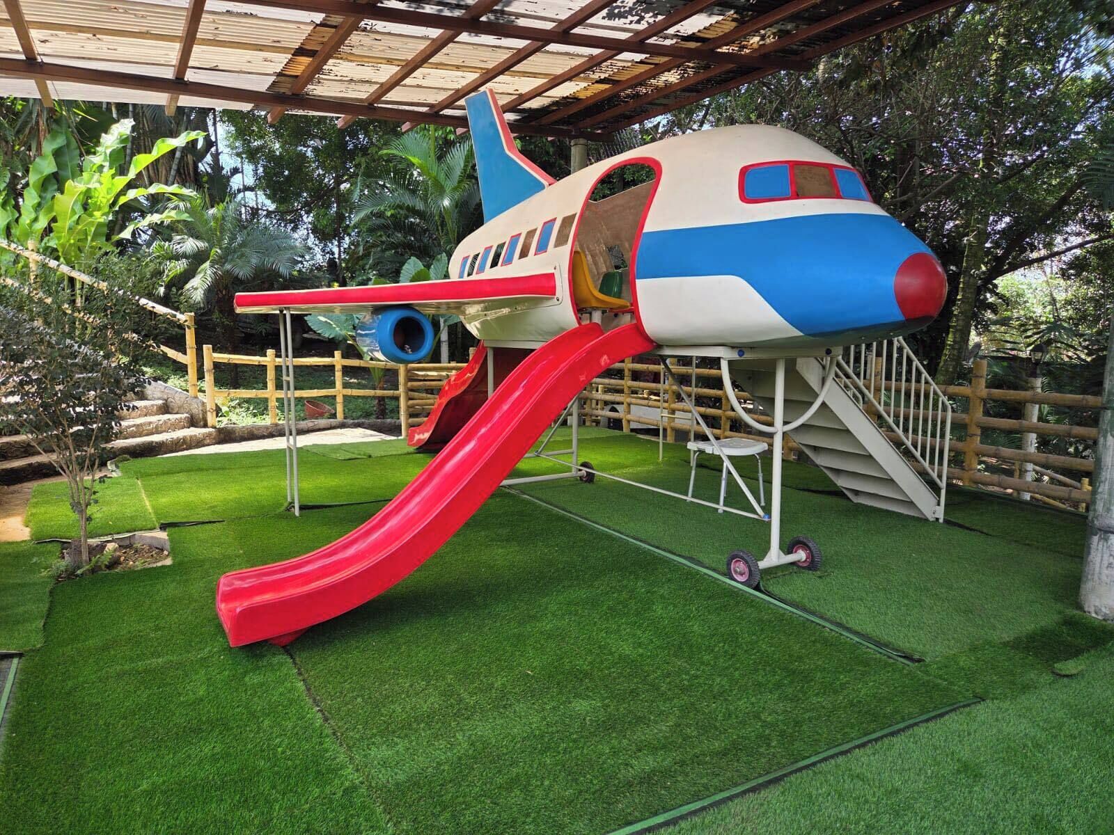 A playground with a slide and climbing area, surrounded by green grass and trees.