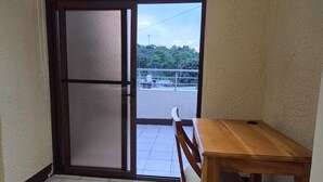 A room with a sliding glass door leading to an outdoor balcony with a railing and greenery. A wooden table and chair are positioned near the door.
