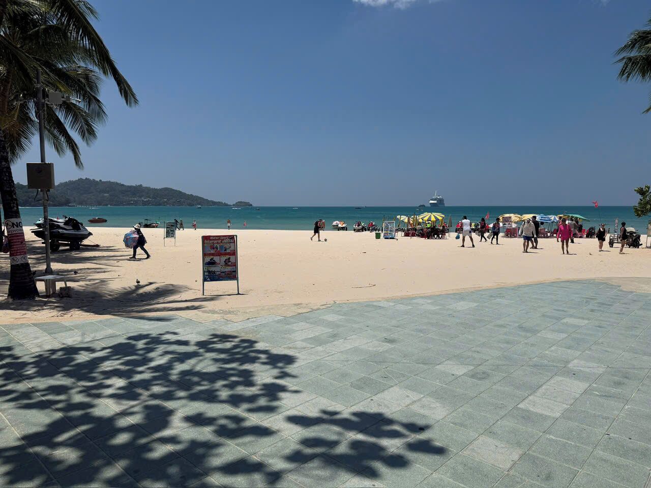 Una playa con un paseo pavimentado, palmeras y un cartel.