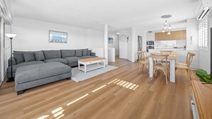 Un salon moderne avec un canapé d’angle gris, une table basse en bois et un coin repas comprenant une table blanche et des chaises en bois.