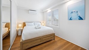 Une chambre d’hôtel avec un lit en bois, du linge de lit blanc, une table de chevet avec une lampe, un climatiseur fixé au mur et une grande fenêtre avec des stores.