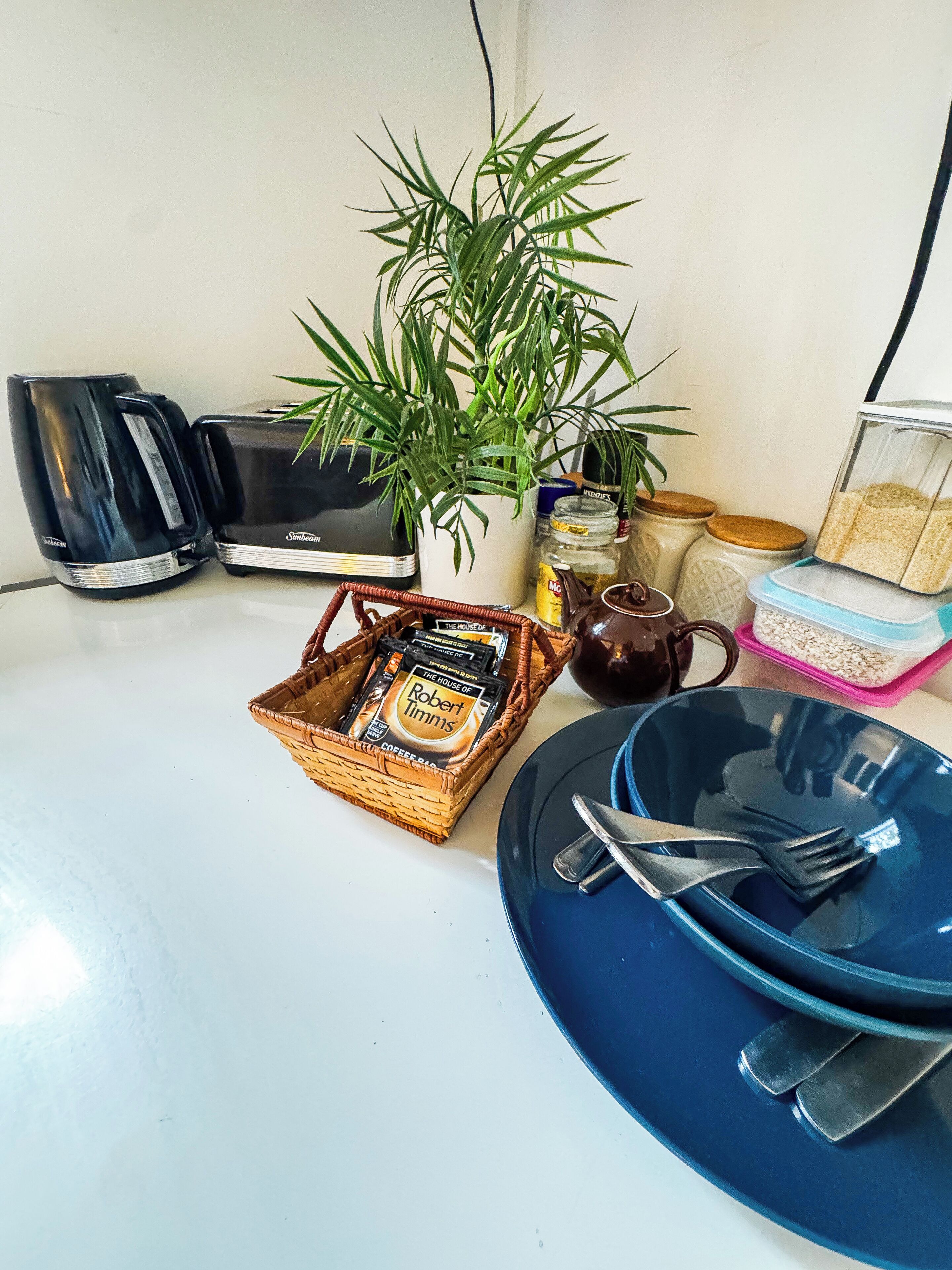 A kitchen counter with a toaster, a coffee maker, a basket of coffee pods, a plant, and various kitchen items.