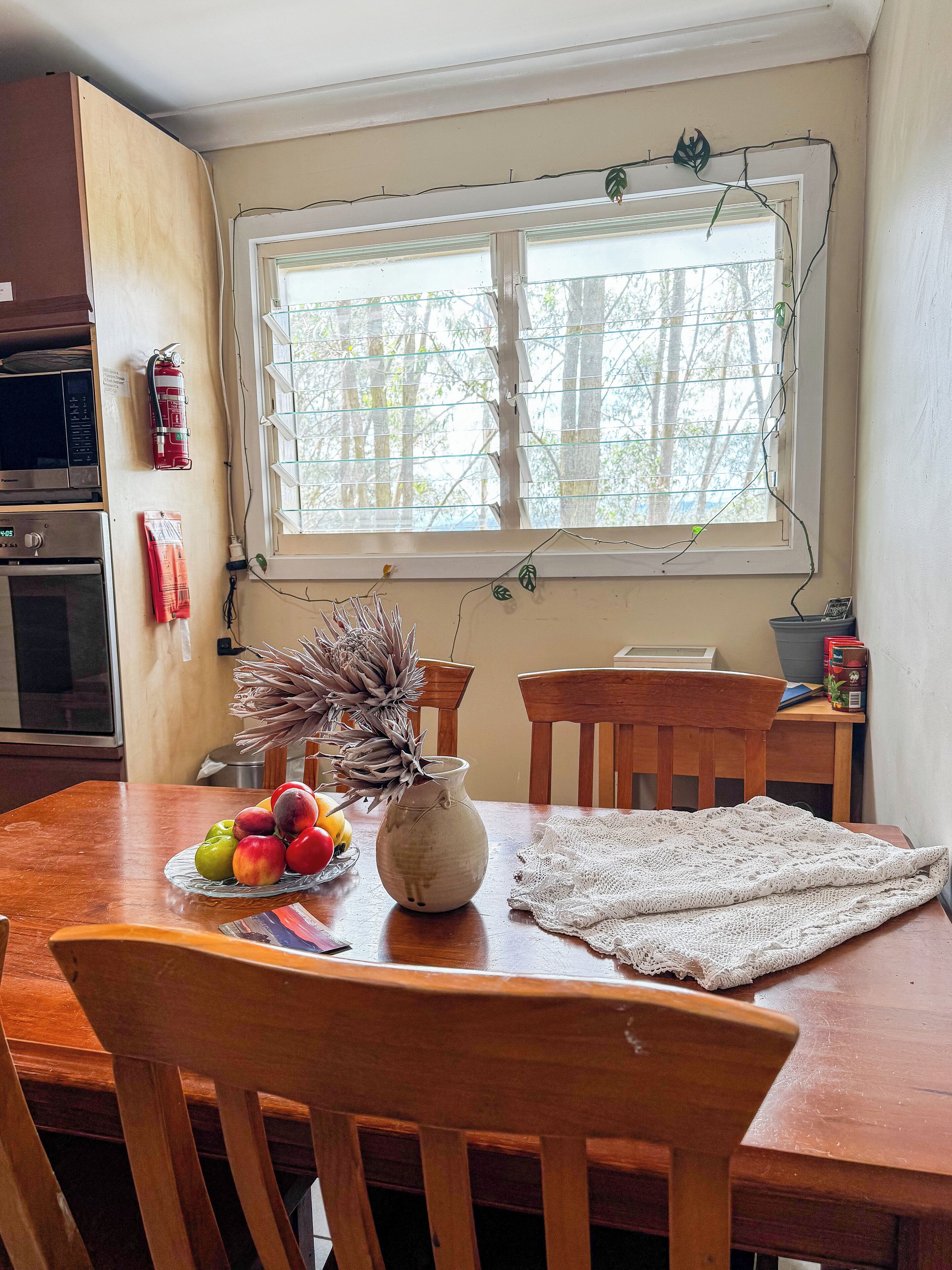 Uma sala de jantar com uma mesa de madeira, cadeiras, um vaso com plantas secas e uma fruteira.