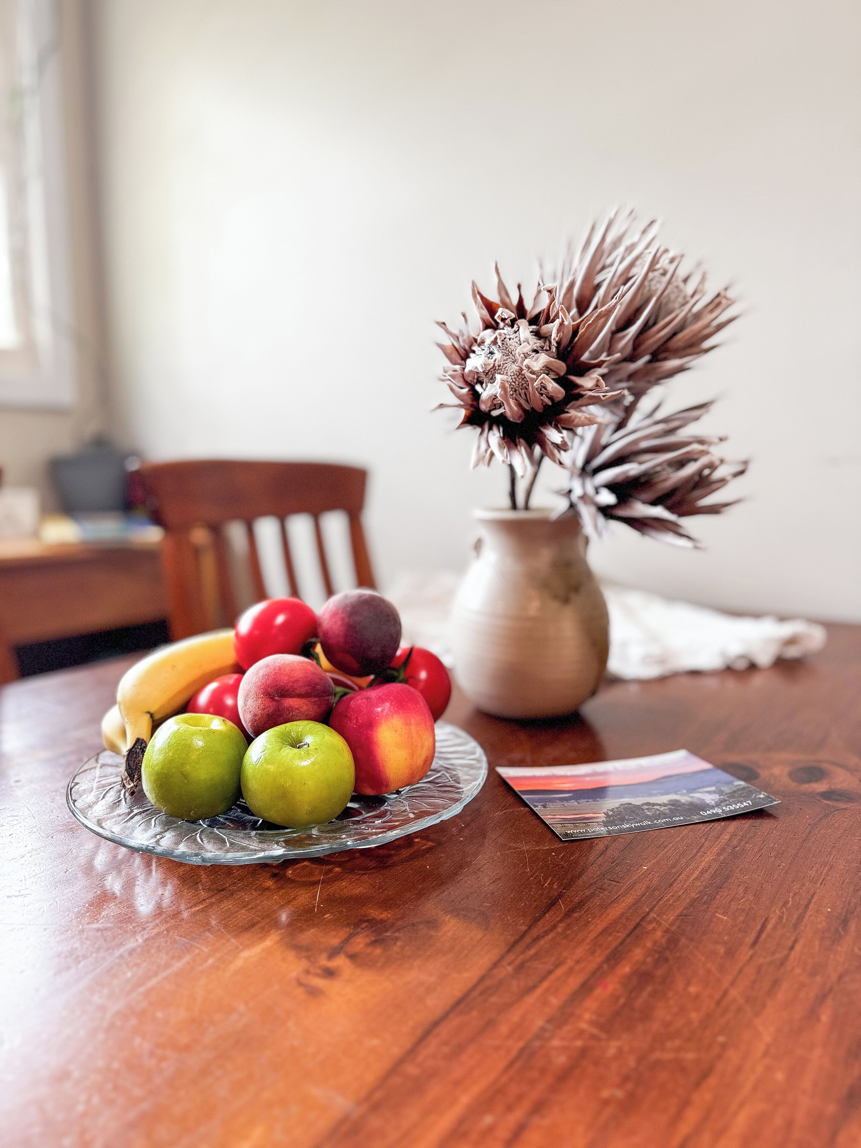 A wooden table with a fruit bowl containing apples, bananas, and peaches, a vase with dried flowers, and a postcard.