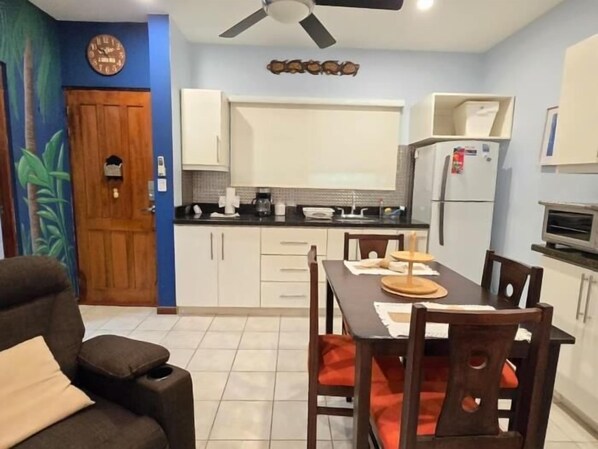 A compact kitchen with white cabinets, a dining table with chairs, a ceiling fan, and a clock on the wall.