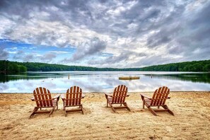 Une plage avec quatre chaises Adirondack en bois, disposées face à un lac paisible, le tout entouré d’une zone boisée.