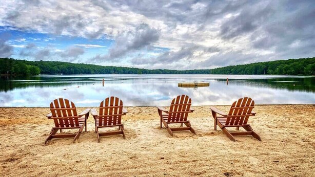A beach with four wooden Adirondack chairs facing a calm lake, surrounded by a forested area.