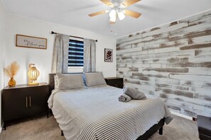 A bedroom with a bed, a nightstand, a lamp, a ceiling fan, and a wall with a wood-like texture. - Tranquil Trails Cottage Fire Pit Cozy 3BDR (Big Bass Lake)