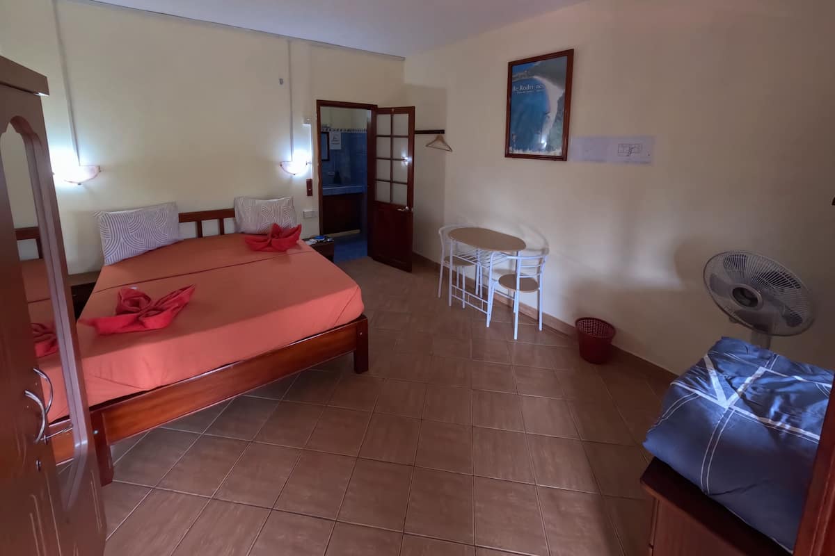 A room with a bed, a small table, a chair, a fan, and a door leading to a bathroom.