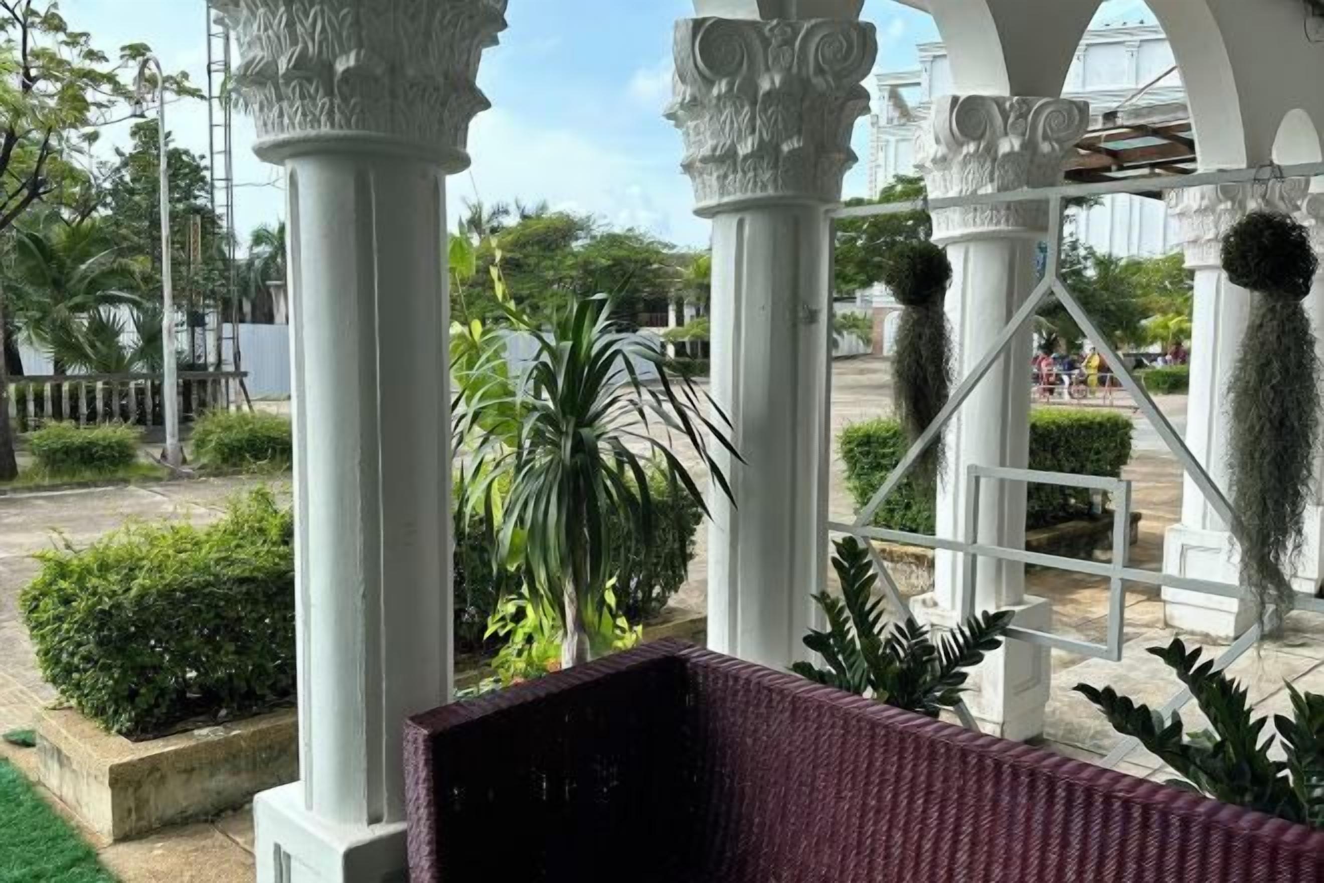 A covered outdoor area with white columns, a wicker bench, and greenery.