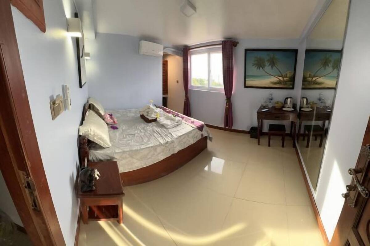 Une chambre à coucher avec un lit, une petite table, une chaise, une fenêtre avec des rideaux et un tableau au mur.