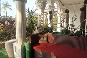 A covered outdoor seating area with a wicker sofa, potted plants, and a pergola structure. - Hawaii Lounge (Sihanoukville)