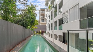 A narrow swimming pool flanked by a wooden fence and a modern building with balconies and large windows. - 2-bed Getaway With Shared Pool & BBQ Access (Brisbane)