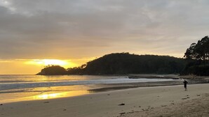 Ein Strand mit einer Person, die im Sand steht, einem Sonnenuntergang im Hintergrund und einem bewaldeten Hügel.