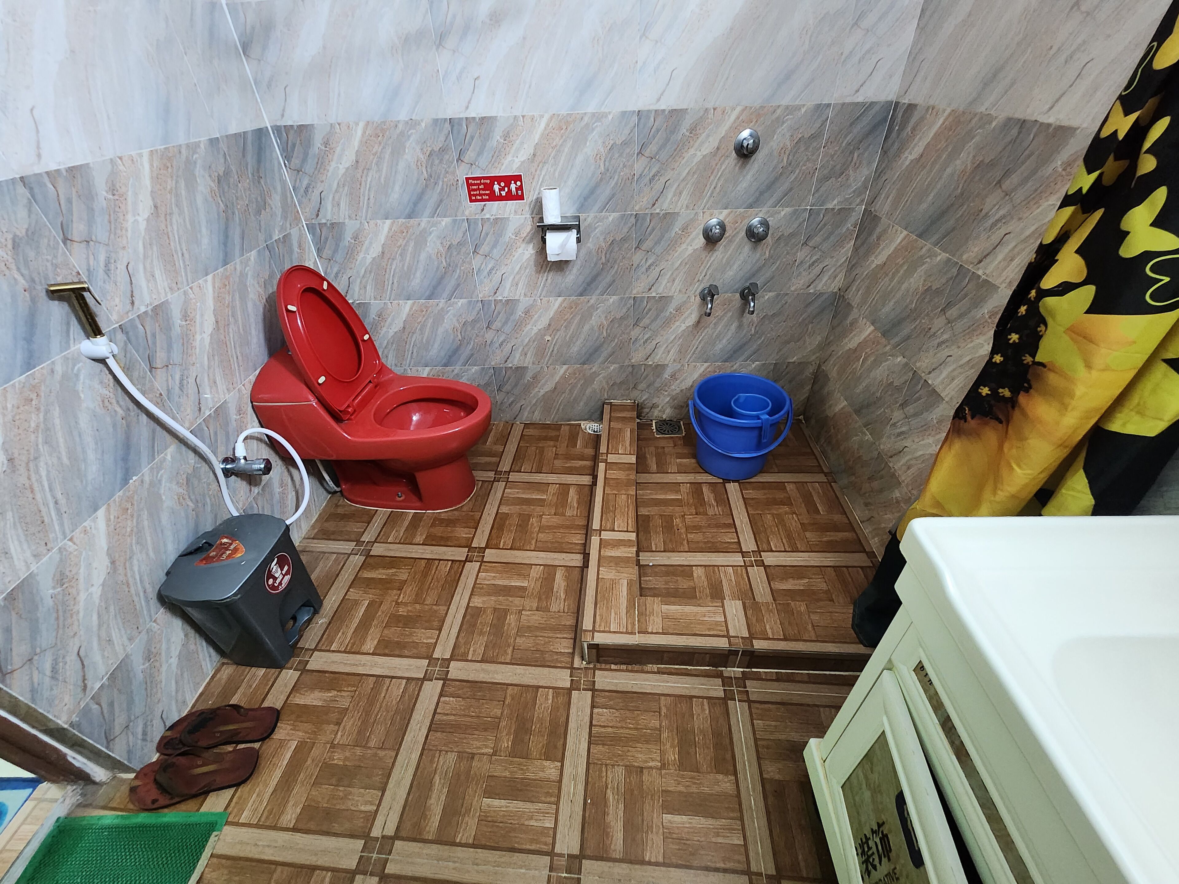 Une salle de bain avec des toilettes rouges, un seau bleu, un lavabo blanc et un rideau de douche.