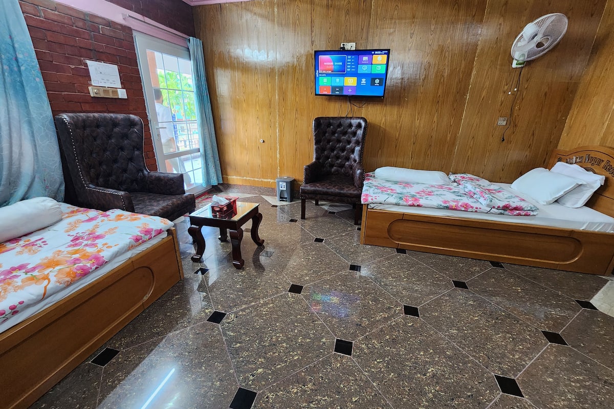 A hotel room with two beds, a wooden floor, a TV on the wall, and a fan.