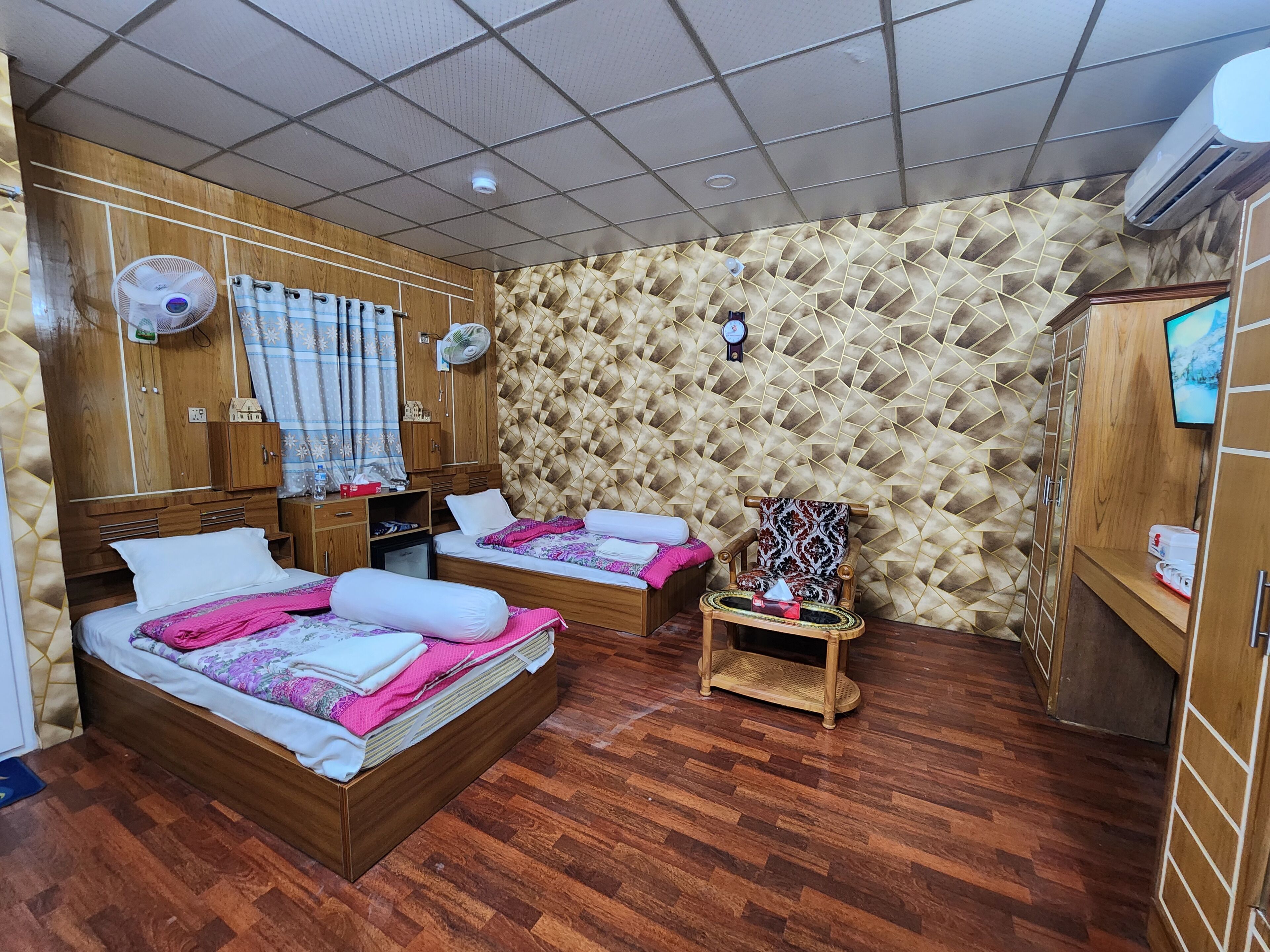 A hotel room with two beds, wooden wardrobe, wall-mounted air conditioner, and a small table with a chair.