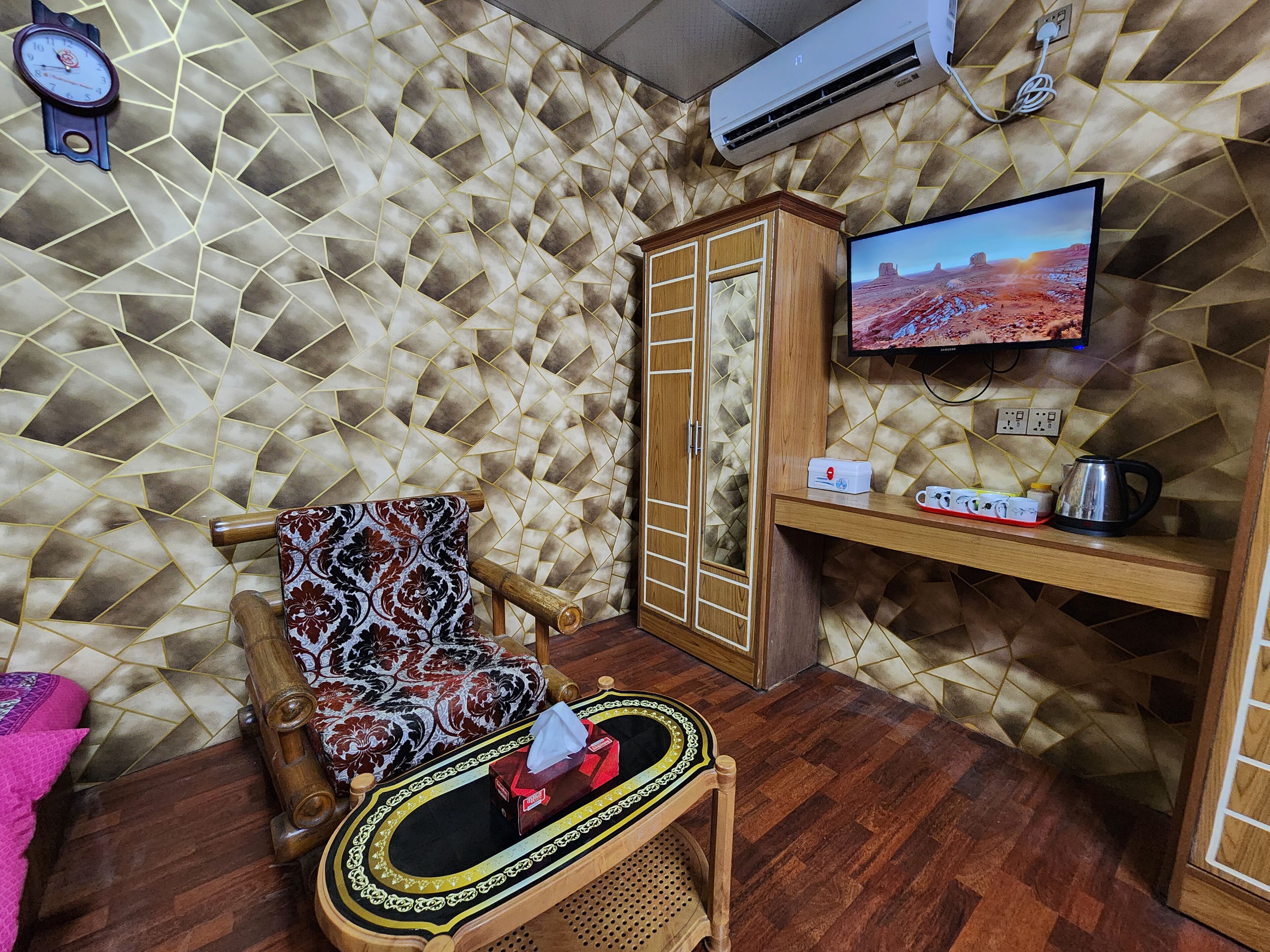 A room with a patterned wall, a wooden wardrobe, a television on a stand, a small table with a tissue box, and a clock on the wall.
