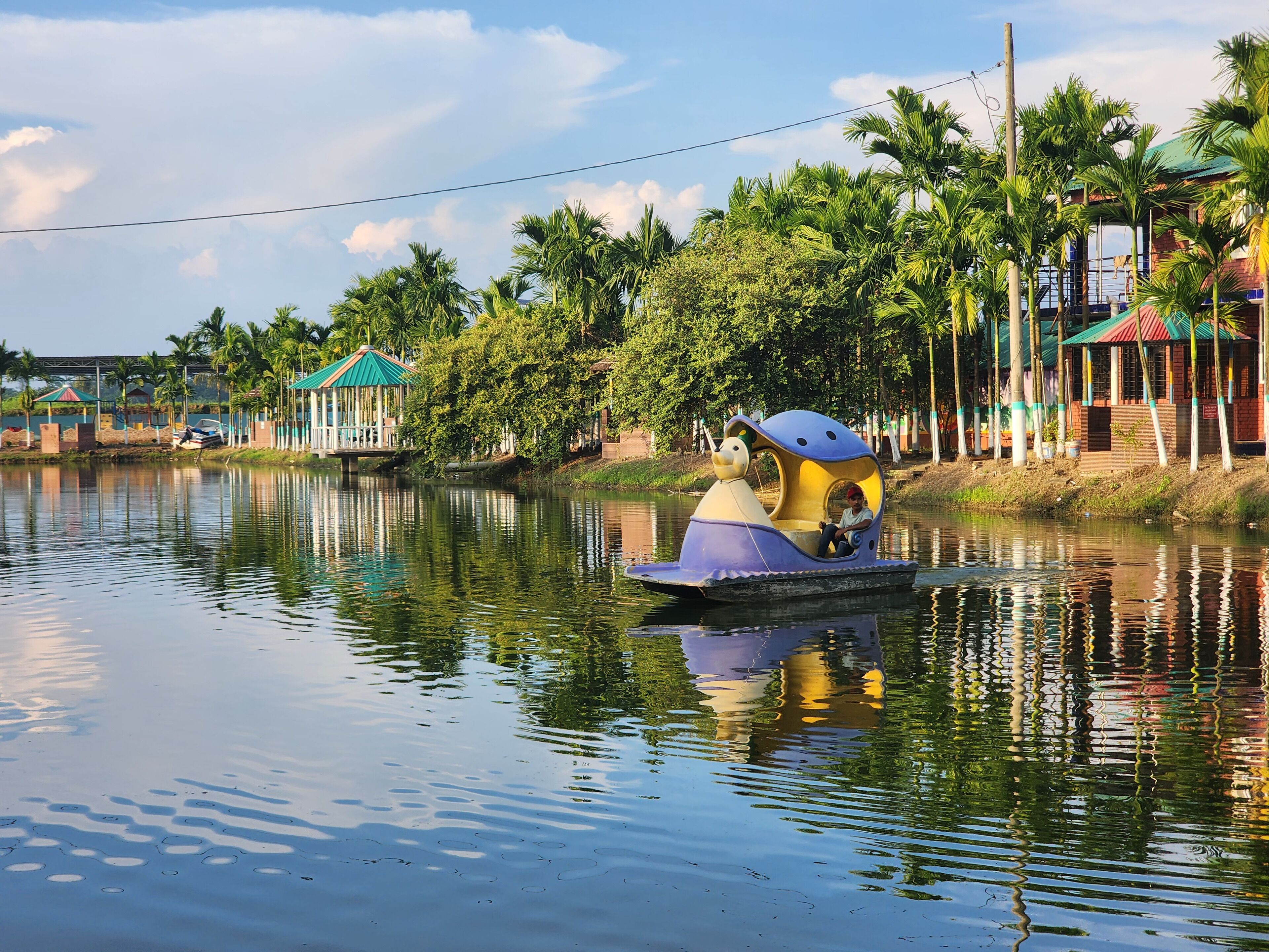 A calm river with a boat carrying a child, surrounded by lush greenery and buildings with colorful roofs.
