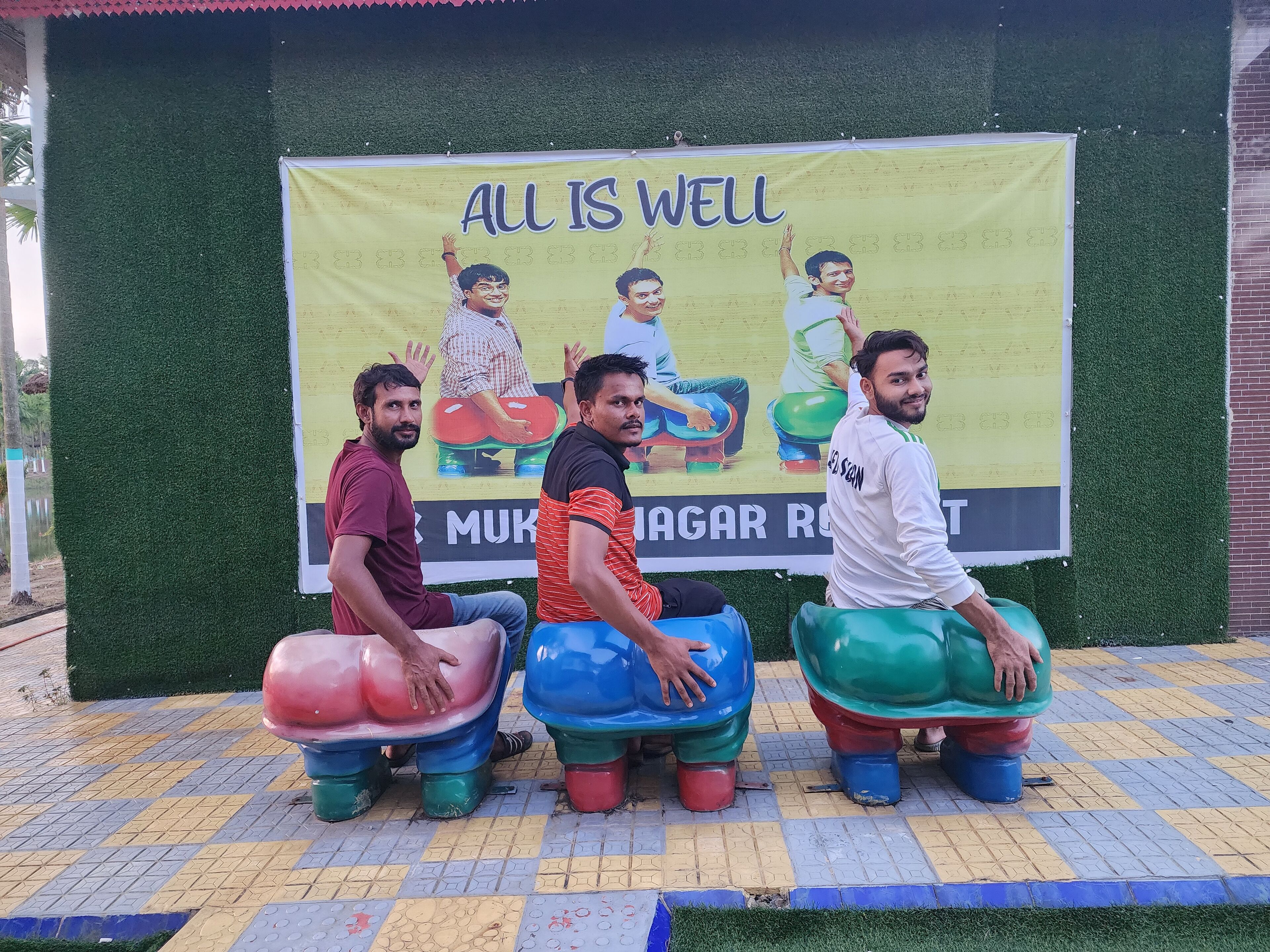 Three men sitting on colorful stools in front of a banner with the text "ALL IS WELL".