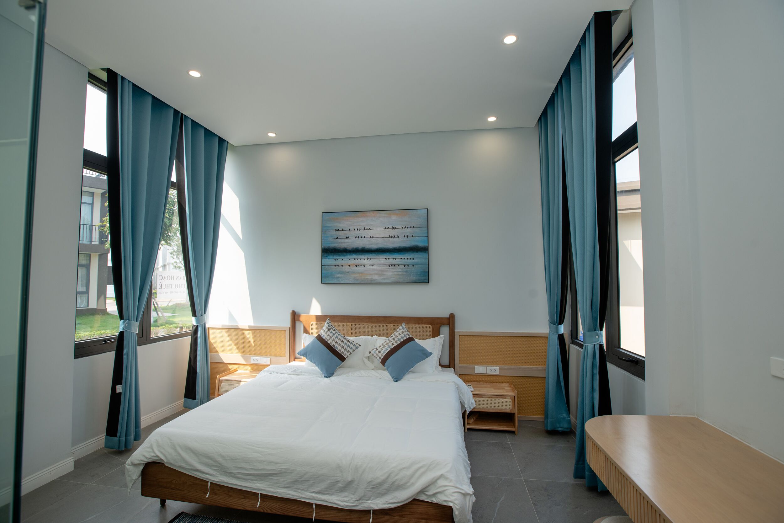 A modern bedroom with a large bed, a wooden headboard, a bedside table, and a view of the outside through a window.