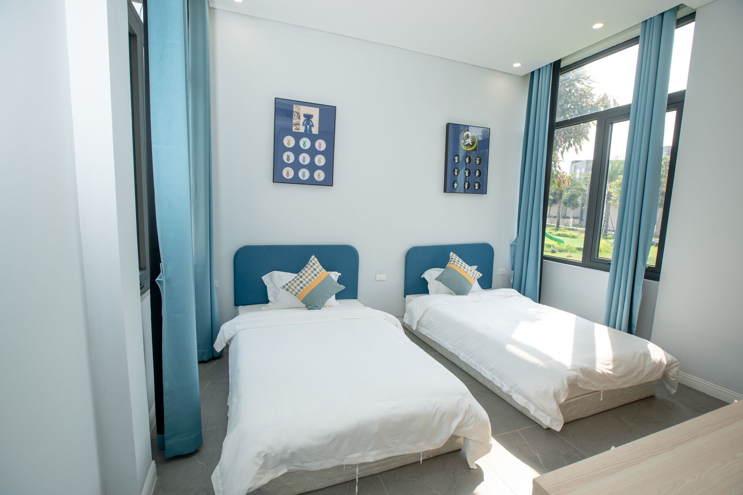 A hotel room with two beds, a large window with curtains, and a wall-mounted control panel.