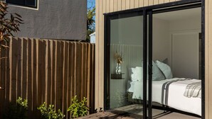 A modern bedroom with a sliding glass door leading to a balcony, a bed with white bedding, and a small bedside table with a vase. - Spacious Central Townhouse – Perfect for All (Christchurch)
