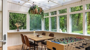 A room with a wooden table, chairs, a foosball table, hanging plants, and large windows with a view of greenery. - Leas Pärla by Interhome (Torup)