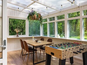 A room with a wooden table, chairs, a foosball table, hanging plants, and large windows with a view of greenery.