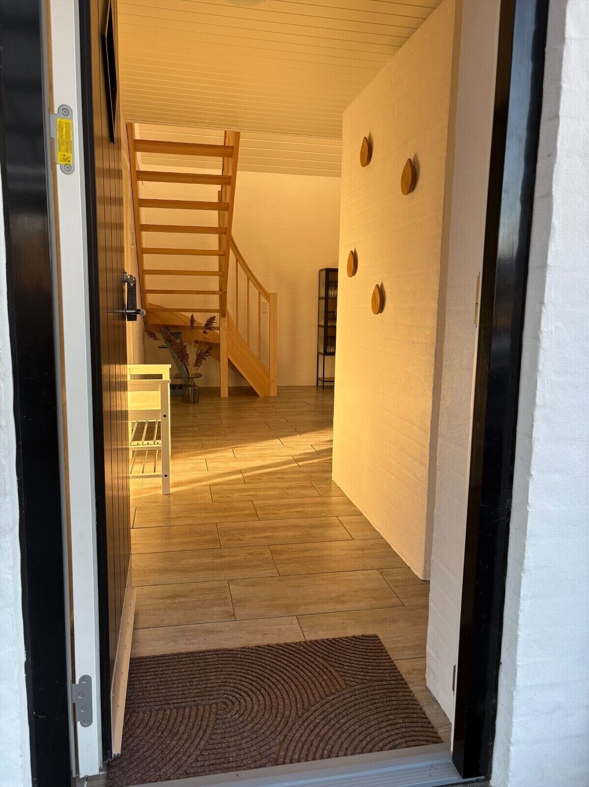 Un escalier en bois menant à l’étage supérieur, un mur blanc orné de trois motifs circulaires et un paillasson marron à l’entrée.