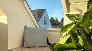 Ein Dachfenster mit Blick auf ein Haus, ein graues Kissen und eine große, belaubte Pflanze.