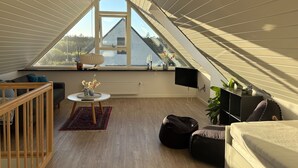 Ein modernes Schlafzimmer im Loft-Stil mit schrägen Wänden, großem Dachfenster, Holzboden, einem Bett, einer Couch, einem Fernseher und einem kleinen Tisch mit Dekoration.