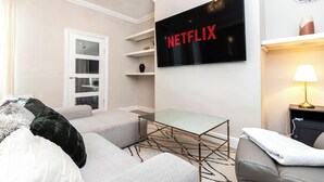 A living room with a glass coffee table, a sofa, a TV mounted on the wall, and a lamp on a side table. - Bright 4-bedroom Maisonette in Gateshead (Gateshead)