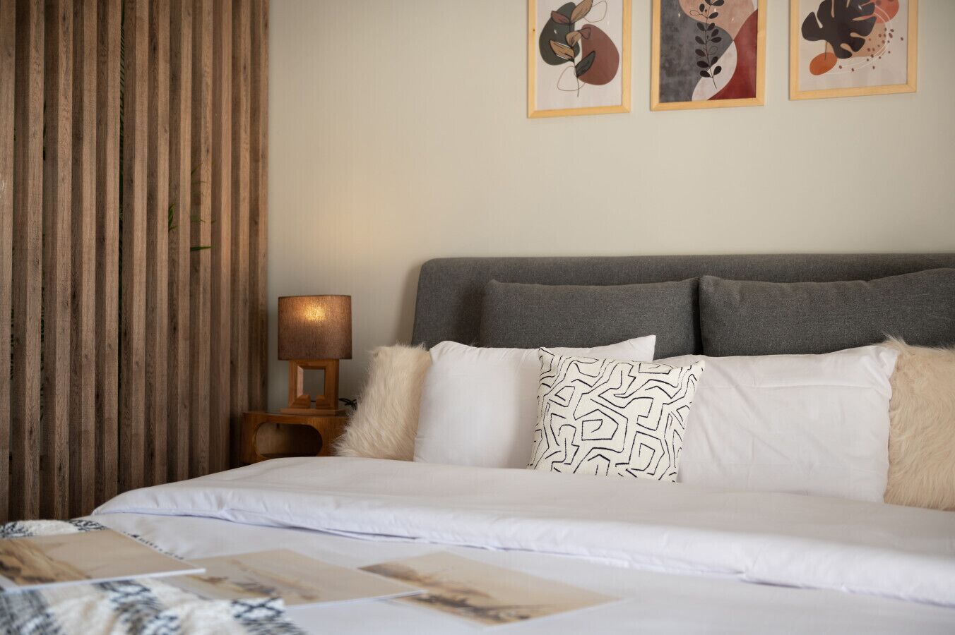A neatly made bed with white linens and patterned pillows, a wooden nightstand with a lamp, and abstract artwork on the wall.
