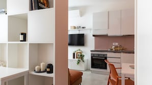 A modern kitchen with white cabinets, a black refrigerator, and a dining area with a table and chairs. - 7Studios - 4 orange (Milano)
